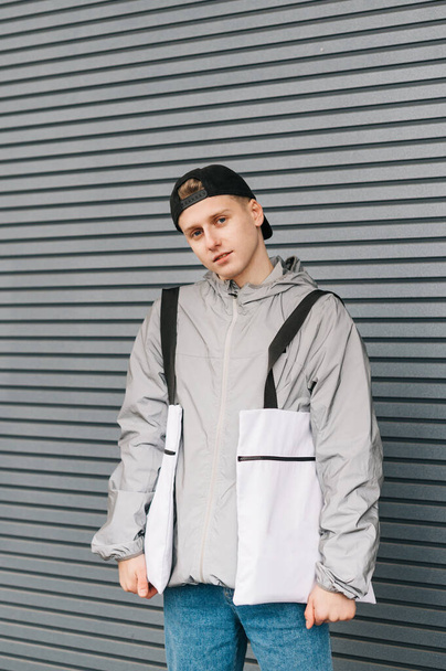 Portrait of a positive young man with 2 eco shopping bags in his hands on a background of a gray wall, looking into the camera with a smile on his face, wearing stylish casual street clothes. Vertical - Fotó, kép