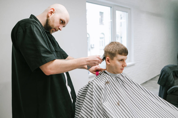Kalju ammattilaisparturi leikkaa asiakkaan hiukset klipperi kädessä, katsoo nuoren miehen päätä. Nuori mies viiltää parturiaan. Parturikonsepti
. - Valokuva, kuva