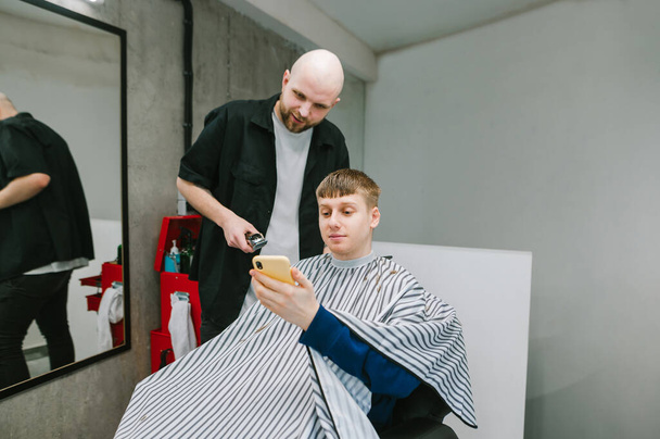 Πελάτης κάθεται στην καρέκλα ενός κομμωτή και δείχνει ένα κομμωτήριο ένα παράδειγμα ενός hairstyle σε ένα smartphone, κουρέας κοιτάζει το τηλέφωνο και χαμόγελα.Ο άνθρωπος με ένα smartphone στο χέρι κόβει τα μαλλιά σε ένα κουρείο - Φωτογραφία, εικόνα