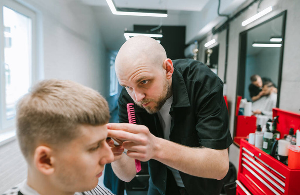A professzionális borbély arra koncentrál, hogy levágja egy fiatal ügyfél haját. Professzionális fodrász csinál egy frizura egy fiatal férfi a fodrászatban, keres egy komoly arc - Fotó, kép