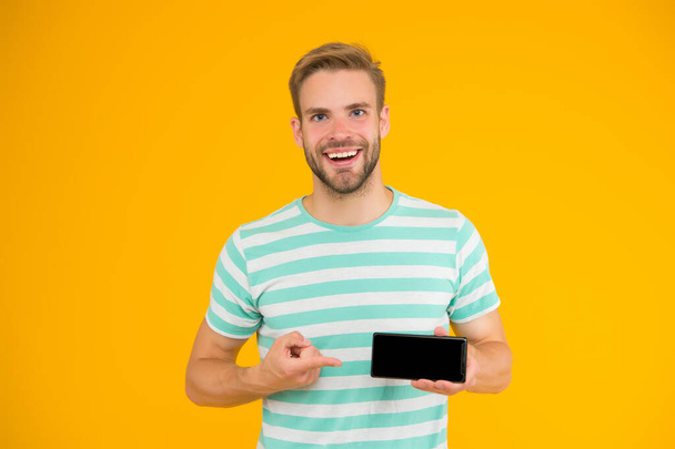 Best phone available for you. Happy guy pointing at smartphone yellow background. Pointing for advertising. Index finger pointing. Pointing gesture. Top pick. It will actually make your life easier - Photo, image