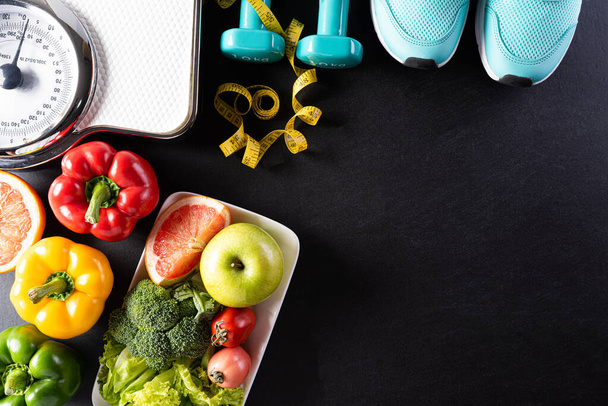 Всемирный день здоровья, здравоохранение и медицинская концепция. Здоровое питание, включая свежие фрукты, овощи, весы, спортивную обувь, гантели и скотч на черном фоне. - Фото, изображение