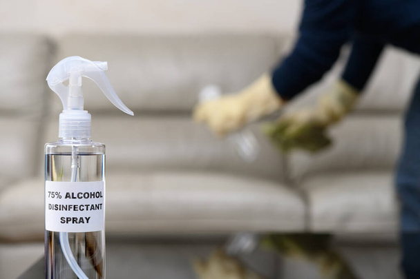 bottle of 75 percent alcohol disinfectant spray with a man disinfecting and cleaning a sofa at background - Photo, Image