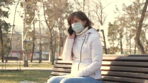 koncepcja zdrowia i bezpieczeństwa. Młoda kobieta w medycznej masce ochronnej siedzi ze smartfonem na ulicy miejskiej w Europie. Ochrona przed koronawirusami. turyści na ulicy nosić maskę ochronną przed wirusami. - Materiał filmowy, wideo