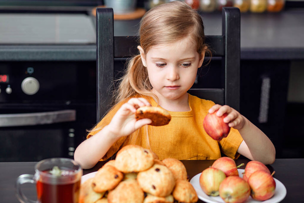 Alimentos saludables y dañinos durante la cuarentena, el autoaislamiento. La pequeña rubia elige tomar una galleta malsana o Apple
.  - Foto, Imagen