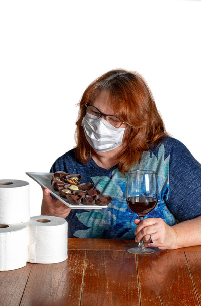 Eine Frau trägt eine Maske während der Haussperre, aber sie kommt mit wichtigen Dingen wie Wein, Schokolade und Toilettenpapier durch. - Foto, Bild