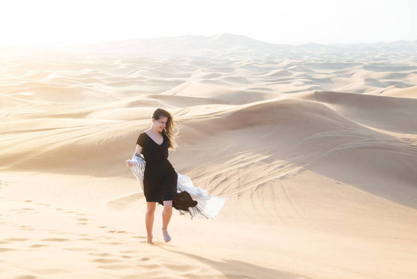 A girl in a black long dress walks through the desert Caucasian young beautiful girl alone is in a hot waterless desert. Runs along sandy mountains and dunes - Photo, image