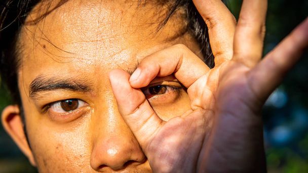 Zblízka pohled na portrét mužských očí vidí skrz mezery v prstech s přirozeným ranním svitem a stíny na tváři. - Fotografie, Obrázek