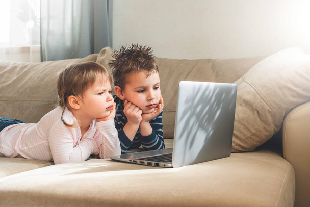 Dzieci uczą się w domu korzystając z lekcji online na laptopie na sofie. Dzieci oglądają kreskówki online. Kwarantanna w domu. Kształcenie na odległość, edukacja online dla dzieci. - Zdjęcie, obraz