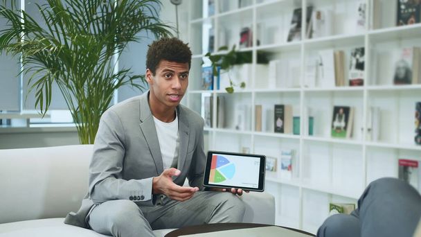 Black formal man making presentation with tablet. Young African-American man in suit sitting in office on sofa with anonymous colleague and making presentation with tablet. - Photo, Image