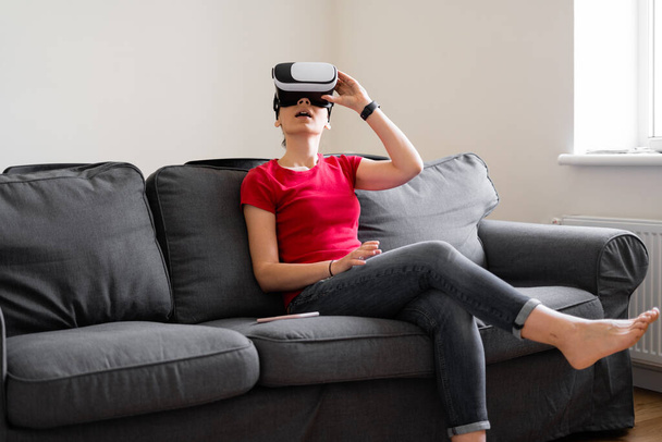 Young woman with VR set sitting on the couch in living room at home. VIrtual reality travel, shopping, learning - Valokuva, kuva