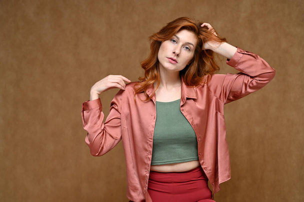 Horizontal portrait of a pretty woman with smooth emotions with long red hair posing in a pink blouse. Photo taken in the studio on a beige background. - 写真・画像