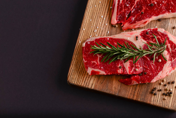Fresh raw beef steak on wooden cutting board over black background with spices, top view. - Photo, Image