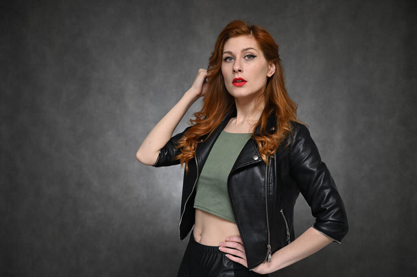 Photo of a red-haired young woman in a black jacket and green t-shirt. Pretty Model with great makeup posing on a gray background in the studio. - Valokuva, kuva