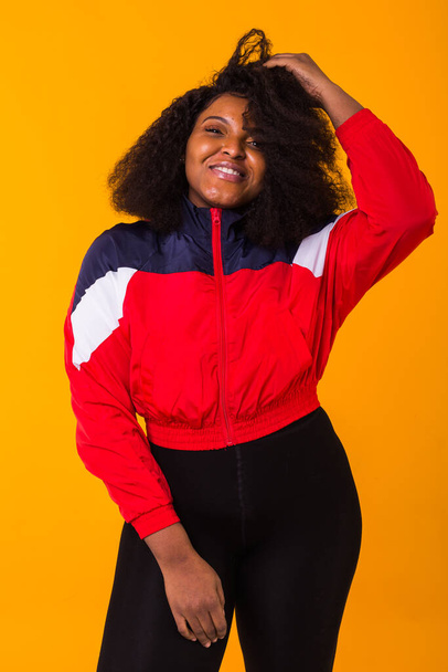 Αστεία κατσαρομάλλη Αφροαμερικανή, ντυμένη με το κόκκινο αθλητικό μπουφάν στο στούντιο σε κίτρινο φόντο. Μόδα και άνθρωποι έννοια. - Φωτογραφία, εικόνα