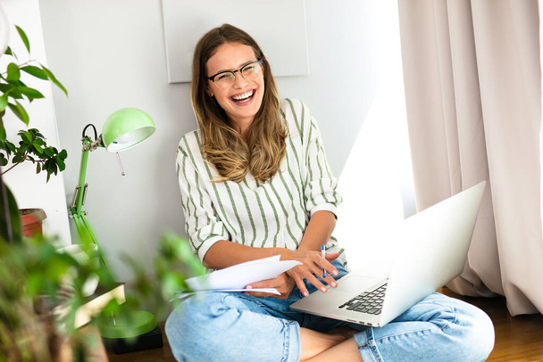 uśmiechnięta kobieta pracująca z domu z laptopem i papperami wokół niej przy jasnym oknie i roślince domowej - Zdjęcie, obraz