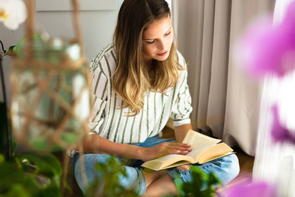 vrouw lezen naast een helder raam, ontspannen en chillen tijdens de uitbraakperiode. Thuisactiviteit voor gezonde geest - Foto, afbeelding