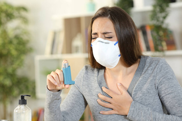 Woman with coronavirus symptoms wearing protective mask holding inhaler suffocating touching chest at home - Foto, Bild