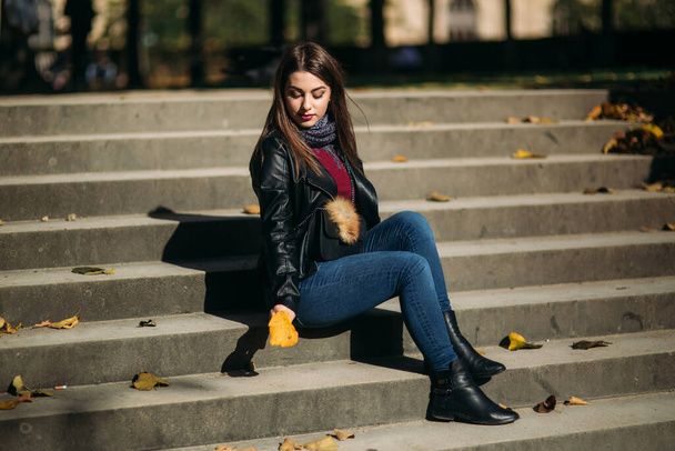 Stylish beautiful lady in a black leather jacket with a black bag and sweatshirt. Female sits on stairs - Photo, Image