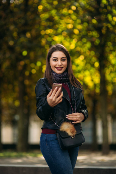 Stylish beautiful lady in a black leather jacket with a black bag and burgundy blouse. Attractive young woman use phone - Photo, Image