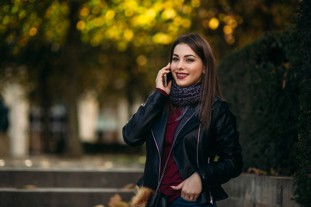 Stylish beautiful lady in a black leather jacket with a black bag and burgundy blouse. Attractive young woman speak by the phone - Photo, Image