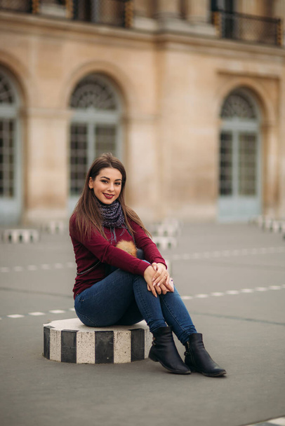 A jovem mulher atraente na blusa borgonha com um lenço cinza senta-se em um estrado listrado. Mulher feliz em Paris
 - Foto, Imagem