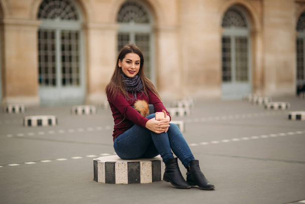 Jolie jeune femme en chemisier bordeaux avec une écharpe grise assise sur un dais rayé. Femme heureuse à Paris
 - Photo, image