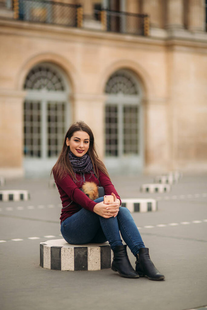 Attractive young womanl in burgundy blouse with a gray scarf sits on a striped dais. Happy woman in Paris - Photo, Image