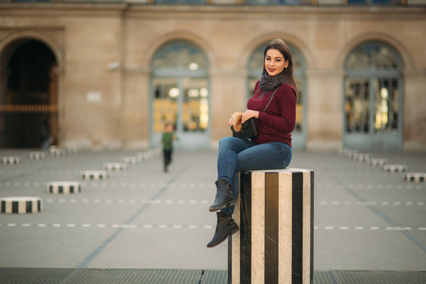 Attractive young womanl in burgundy blouse with a gray scarf sits on a striped dais. Happy woman in Paris - Foto, afbeelding