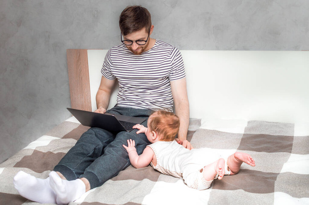 Young man working on a laptop in bed at home and baby beside. Concept Working remotely - Фото, изображение