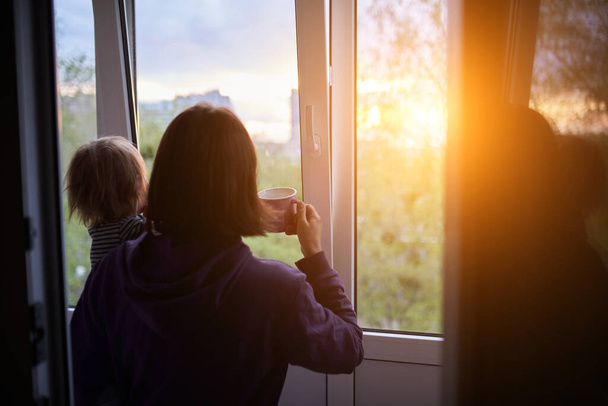 Mother with baby looks out the window at sunset in isolation at home for virus outbreak. Stay home concept. - Foto, Imagen