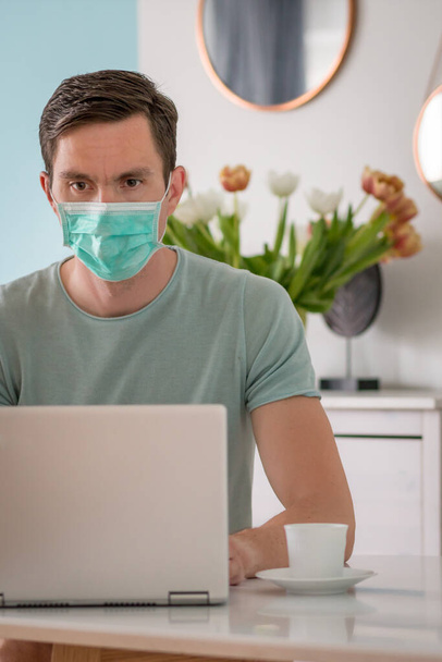 Een man met een ademhalingsmasker ontbijt en werkt op de laptop - Foto, afbeelding