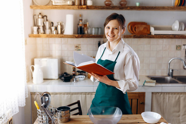 Nuori nainen seisoo keskellä keittiötä reseptikirja käsissään. Hän katsoo kameraan ja hymyilee. Ruoanlaitto kotona käsite, elämäntapa
. - Valokuva, kuva