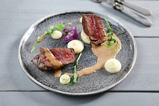 Veal tenderloin filet mignon with root-crop cream in gray plate. Beef slices with sauce, greenery and violet flowers decoration close up. Meat appetizer, restaurant meal, delicious dish - Photo, Image