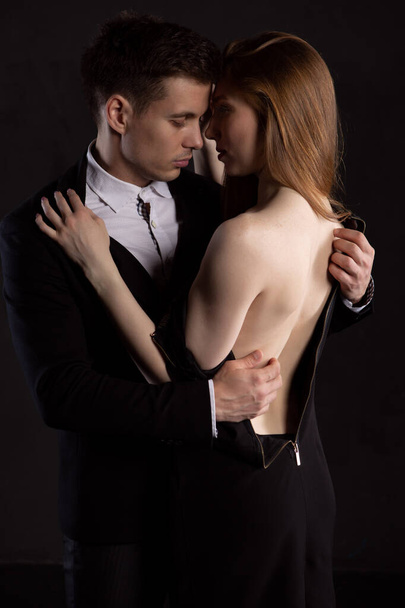 a handsome man is concentrating on taking off the dress of a sexy girl who is gently embracing him and looking at her beautiful Breasts. Fashionable portrait of a couple in love on a dark background - Фото, зображення
