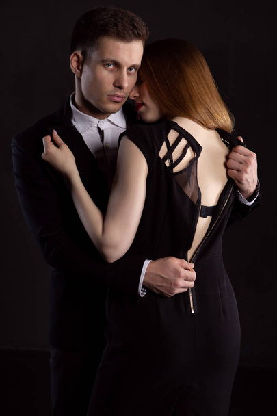 A young man with a stern look unbuttons the elegant dress of the girl who is embracing him. Passionate embrace of a young couple on a dark background - Foto, imagen