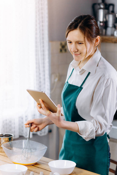 Woman looking to laptop and reading recipe for cooking dishes. Cooking at home concept, lifestyle. Preparing the dough,  baking in the kitchen. - Φωτογραφία, εικόνα