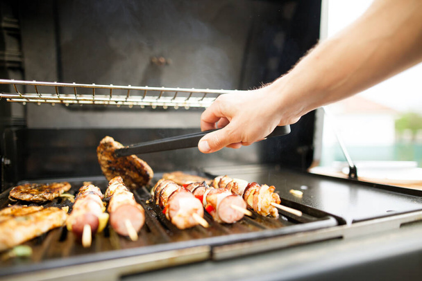 Руки молодого чоловіка перевертають м'ясо виделкою на газовому грилі, концепція барбекю
 - Фото, зображення