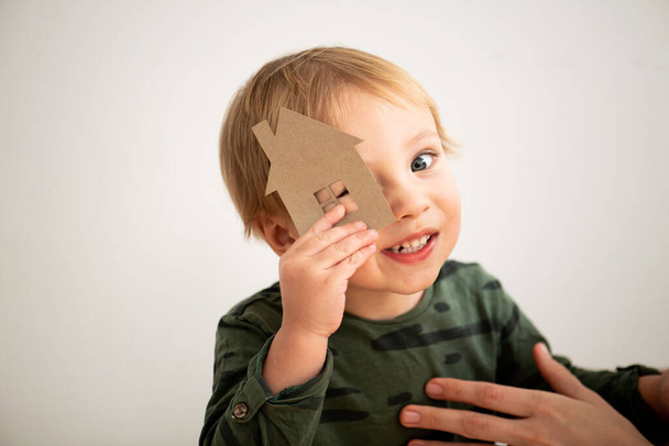 Clumsyblond toddler boy in green long-sleeve trying to look through paper house window. Mother's hand on his chest. Over white wall. - Photo, Image