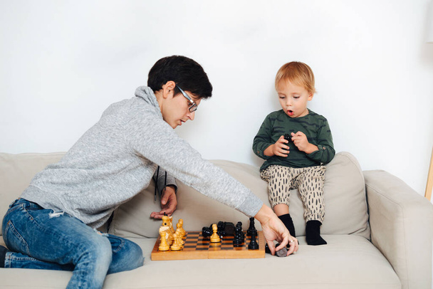 Father teaching his toddler son how to play chess on a sofa at home. They are lining up pieces. Learning how to place them properly. - Foto, immagini