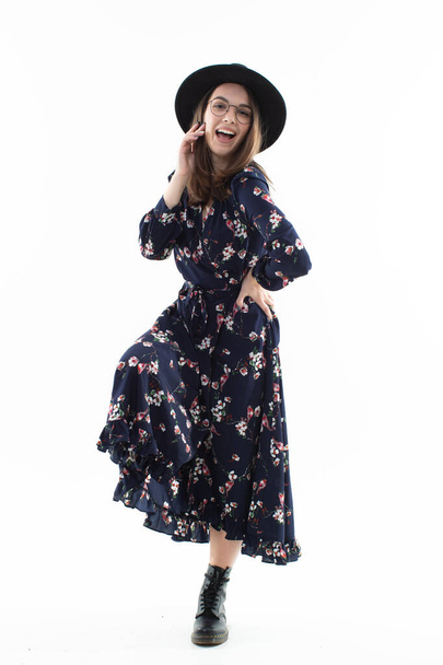 Μια μελαχρινή με μακρύ λουλουδάτο φόρεμα με καπέλο ποζάρει για την κάμερα στο στούντιο. Συναισθηματικό πορτρέτο - Φωτογραφία, εικόνα