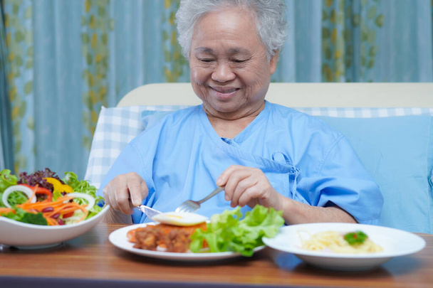 アジアの高齢女性高齢者の女性患者病院でベッドの上に座って空腹ながら希望と幸せと朝食野菜の健康食品を食べる. - 写真・画像