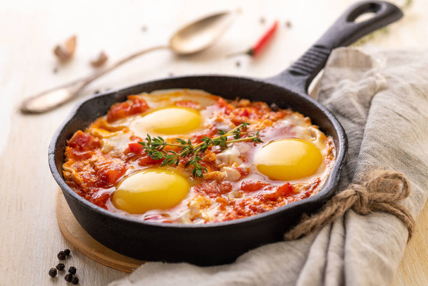 Shakshuka. Fried eggs with tomatoes and sweet pepper and herbs in a serving cast-iron frying pan. Traditional oriental cuisine for breakfast. Close-up. - 写真・画像