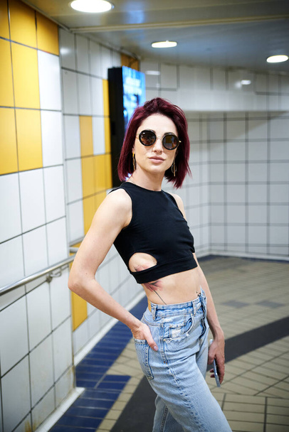 Young woman with burgundy red maroon short hair, wearing black top, light blue jeans, standing in subway station, Three-quarter portrait of pretty girl in black sunglasses at metro station - Photo, image