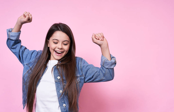 Portrait of a little girl fanny caucasian brunette in casual clothes who is happy to rejoice, dancing, having fun with raised hands on a pink background. Lady wide smile with close eyes, copy space. - Photo, image