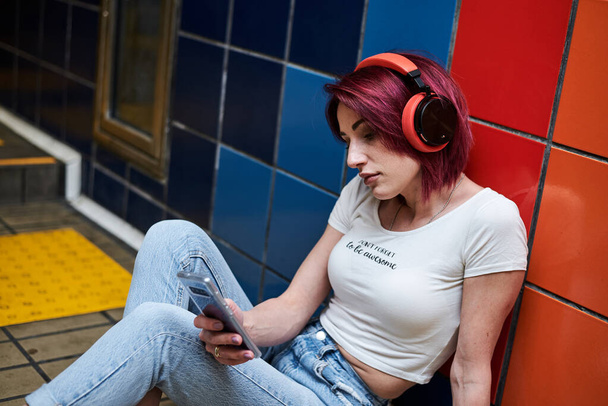 Young woman with burgundy pink maroon hair, in white top and light blue jeans, with red headphones, sitting on floor by colorful wall in subway, Creative portrait of a girl, holding her phone. - Fotografie, Obrázek