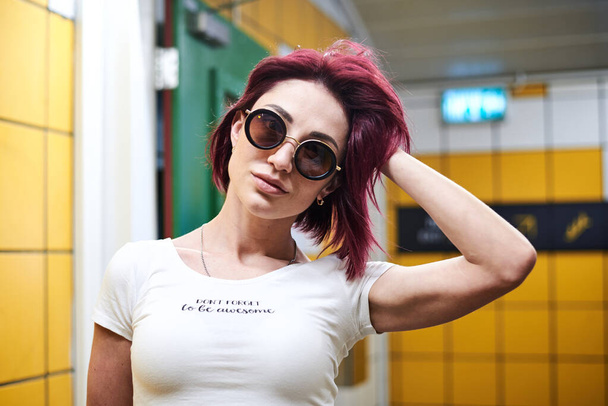 Młoda kobieta z bordowo-różowymi, bordowymi włosami, w białej bluzce i okrągłych czarnych okularach, stojąca w żółtej hali metra, pozująca. Close-up portret ładnej dziewczyny na stacji metra. - Zdjęcie, obraz