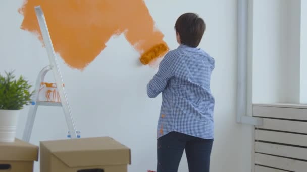 Singleton estilo de vida y concepto de renovación. Concepto de renovación plana. Feliz mujer de mediana edad pintando pared blanca con rodillo de pintura, pintura naranja
 - Metraje, vídeo