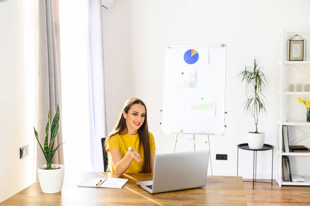 A young woman is contacting an employee via an online video connection. A woman sits indoors and using a laptop and airpods, a flip chart in the background. Remote work - Photo, image