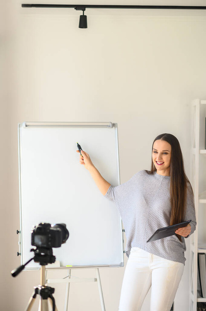 Young woman teacher, tutor in casual wear makes tutorial video on the camera on a tripod indoors . She with tablet in hand points by marker on a flipchart, . The concept of online education, online learning - Photo, Image